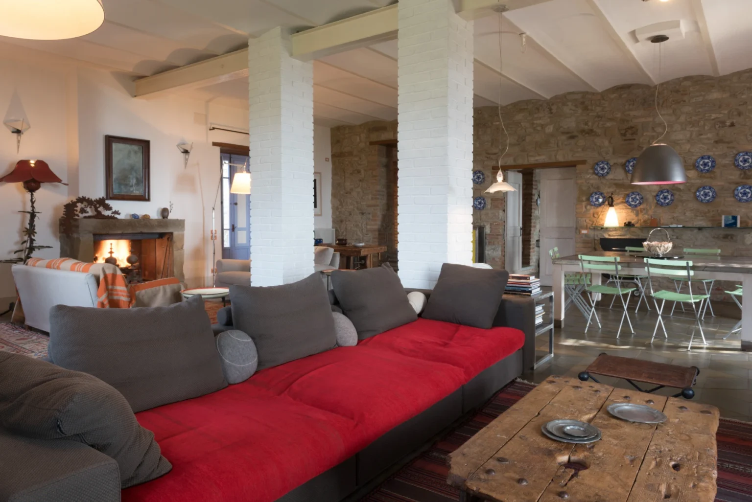 Cozy living room featuring a red sofa, rustic wooden coffee table, and a blend of modern and traditional decor, with an open layout connecting to a dining area and kitchen. Natural light illuminates the space through large windows, enhancing the warm ambiance. | Only on SopranoVillas