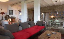 Cozy living room featuring a red sofa, rustic wooden coffee table, and a blend of modern and traditional decor, with an open layout connecting to a dining area and kitchen. Natural light illuminates the space through large windows, enhancing the warm ambiance.