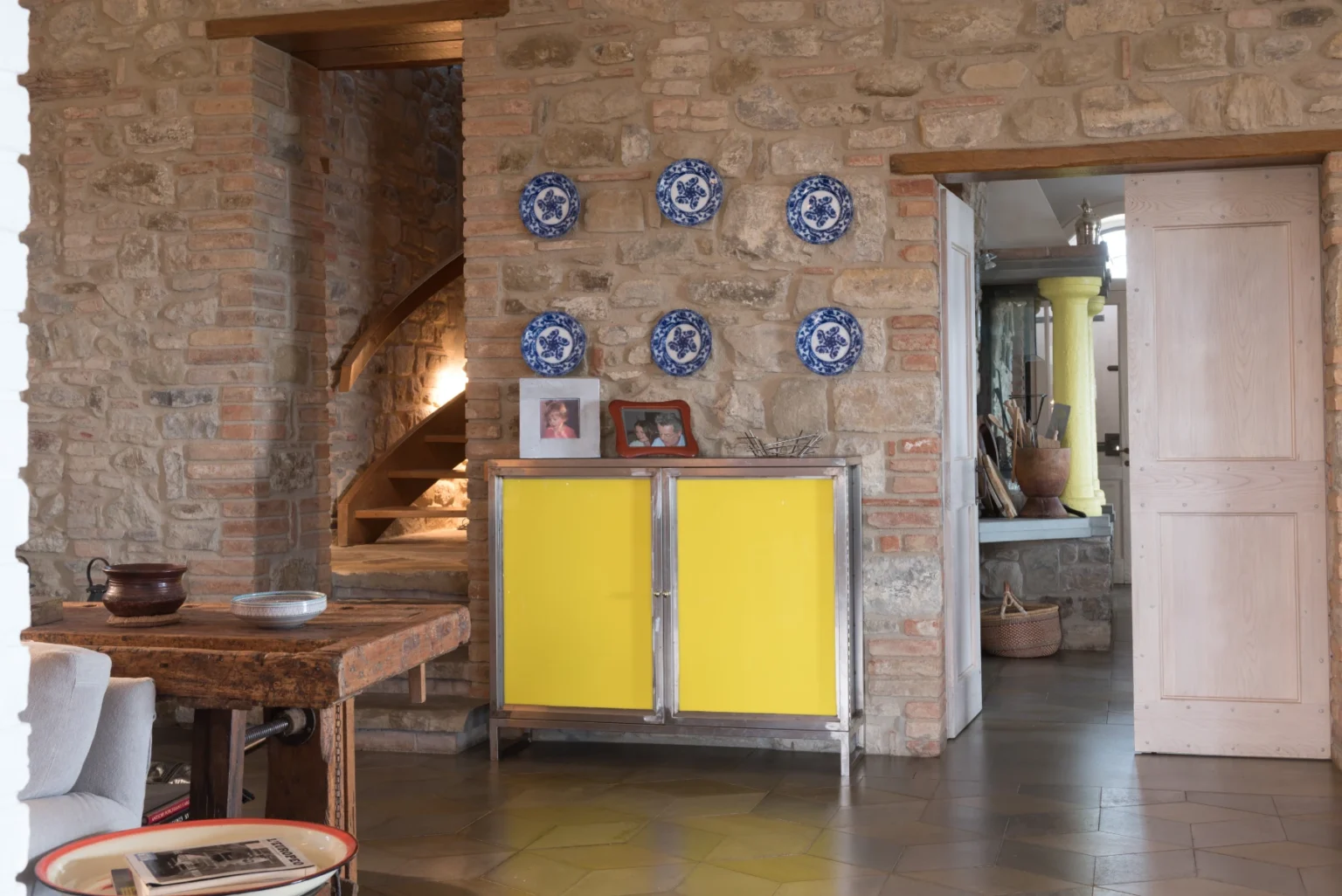 Interior view of a rustic living space featuring exposed stone walls, a wooden dining table, and a vibrant yellow cabinet. Decorative blue and white plates are mounted on the wall, and a doorway leads to another room, enhancing the cozy ambiance. | Only on SopranoVillas