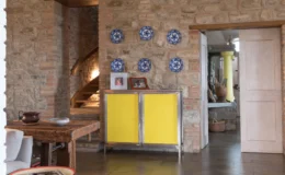 Interior view of a rustic living space featuring exposed stone walls, a wooden dining table, and a vibrant yellow cabinet. Decorative blue and white plates are mounted on the wall, and a doorway leads to another room, enhancing the cozy ambiance.