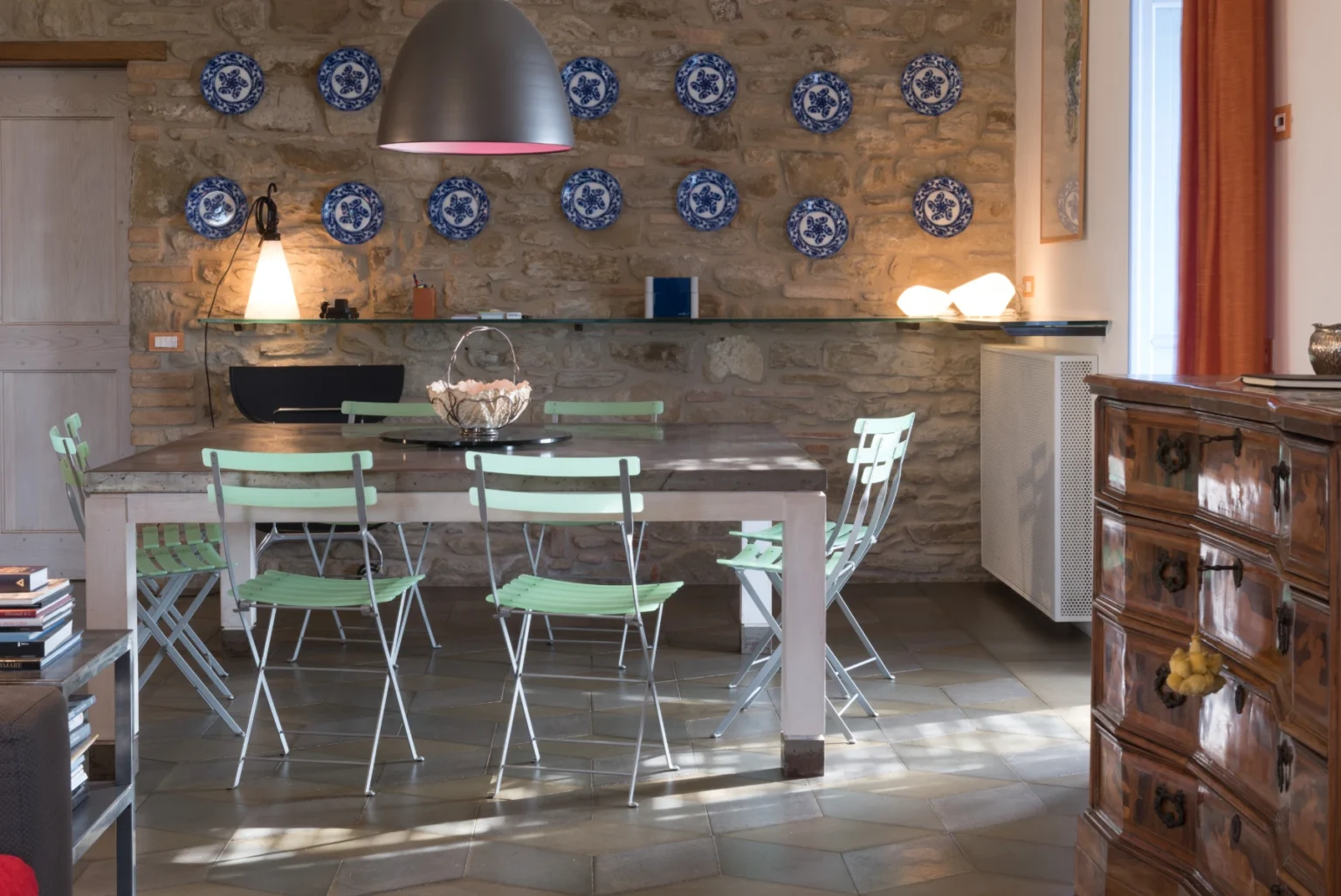 Modern kitchen interior featuring a wooden dining table surrounded by green metal chairs, a stone wall adorned with decorative blue plates, and a stylish pendant light. | Only on SopranoVillas