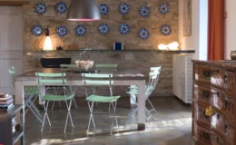 Modern kitchen interior featuring a wooden dining table surrounded by green metal chairs, a stone wall adorned with decorative blue plates, and a stylish pendant light.