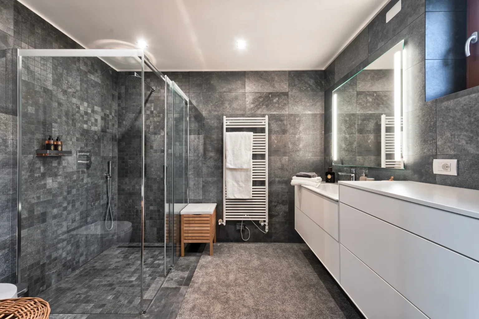 Grey-tiled bathroom with a glass shower enclosure, a long white vanity and illuminated mirrors. | Only on SopranoVillas