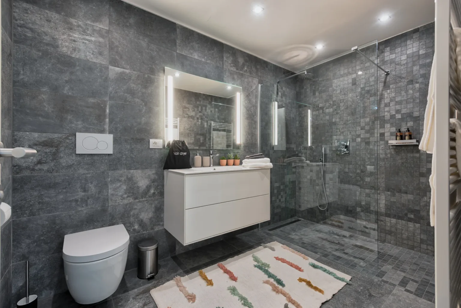 Grey-tiled bathroom with a glass shower enclosure, a white vanity and a wall-hung toilet. | Only on SopranoVillas