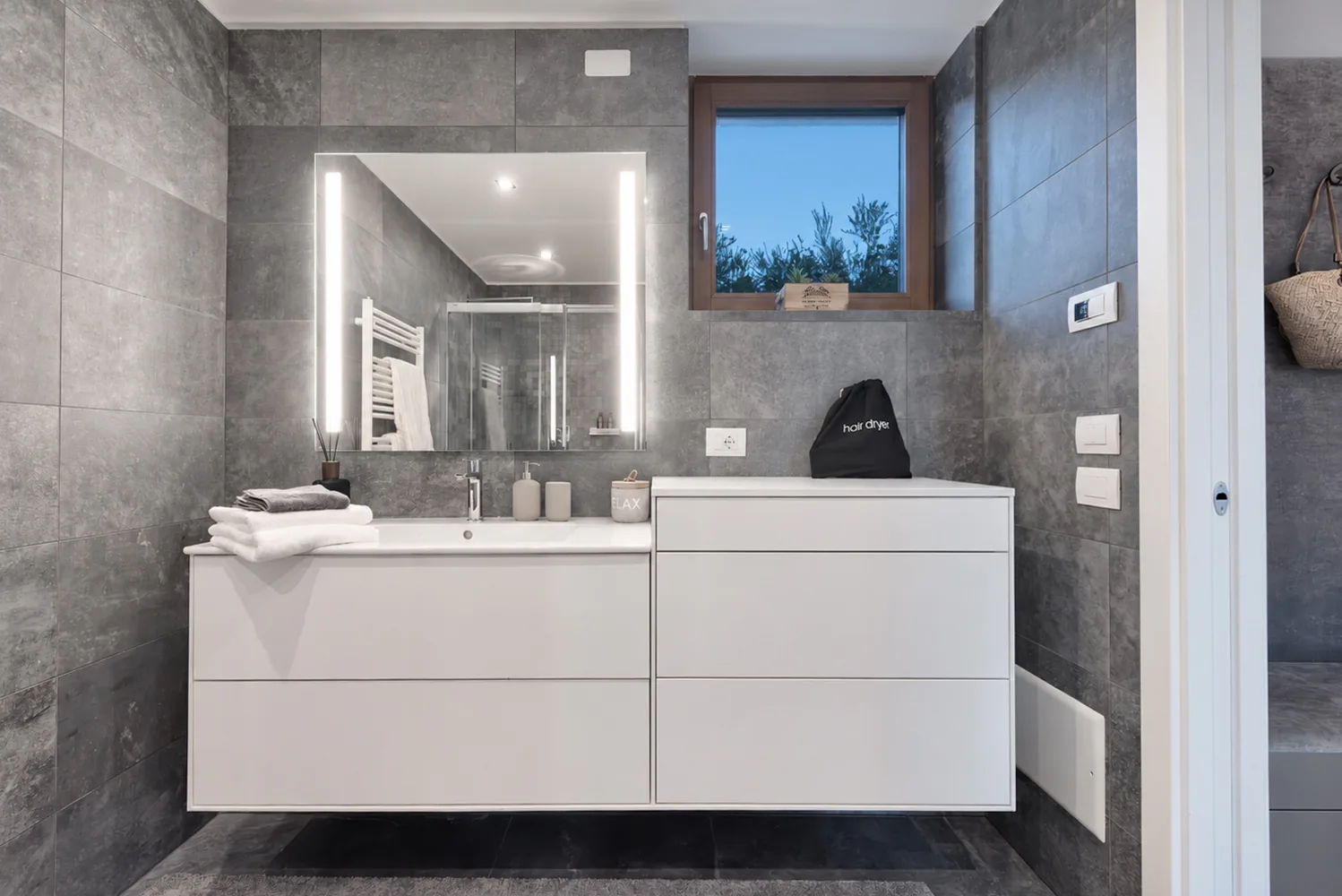 White bathroom vanity beneath an illuminated mirror, beside a window and grey tiled walls. | Only on SopranoVillas