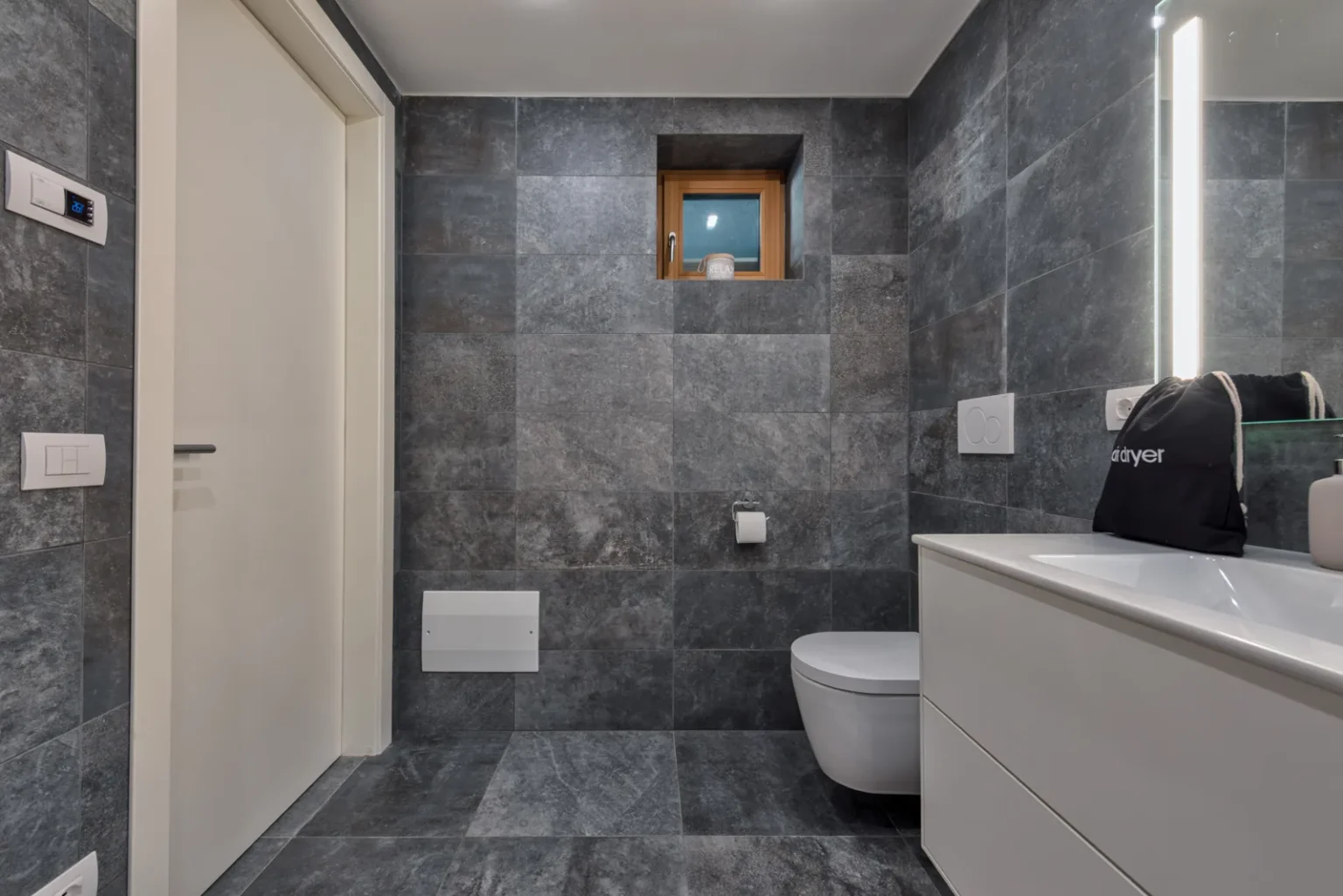 Bathroom with a wall-hung toilet, a white vanity and a small window above the grey tiled wall. | Only on SopranoVillas