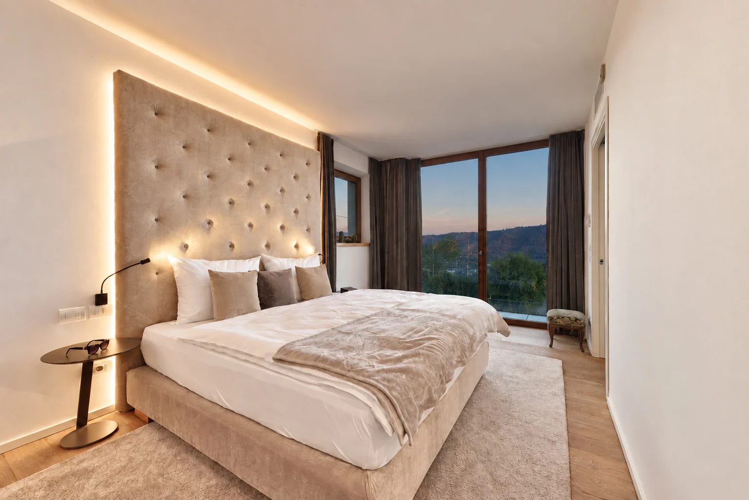 Bedroom with a tall upholstered headboard, warm lighting and a large window overlooking the hills. | Only on SopranoVillas