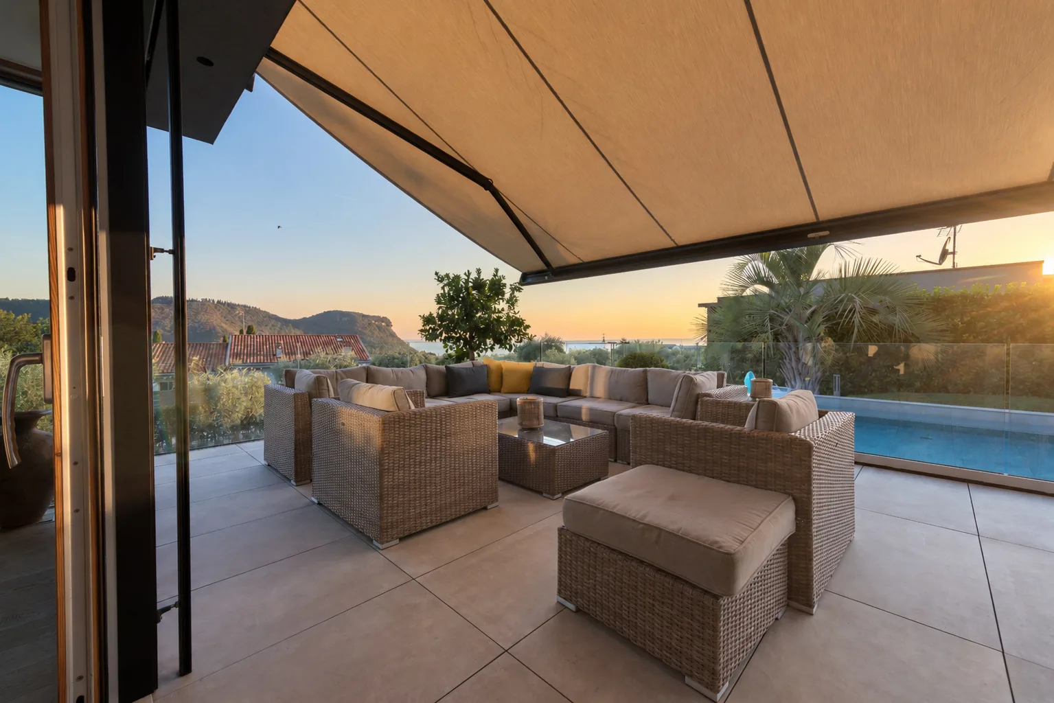 Covered poolside terrace with woven sofas, an armchair and glass railings at sunset. | Only on SopranoVillas