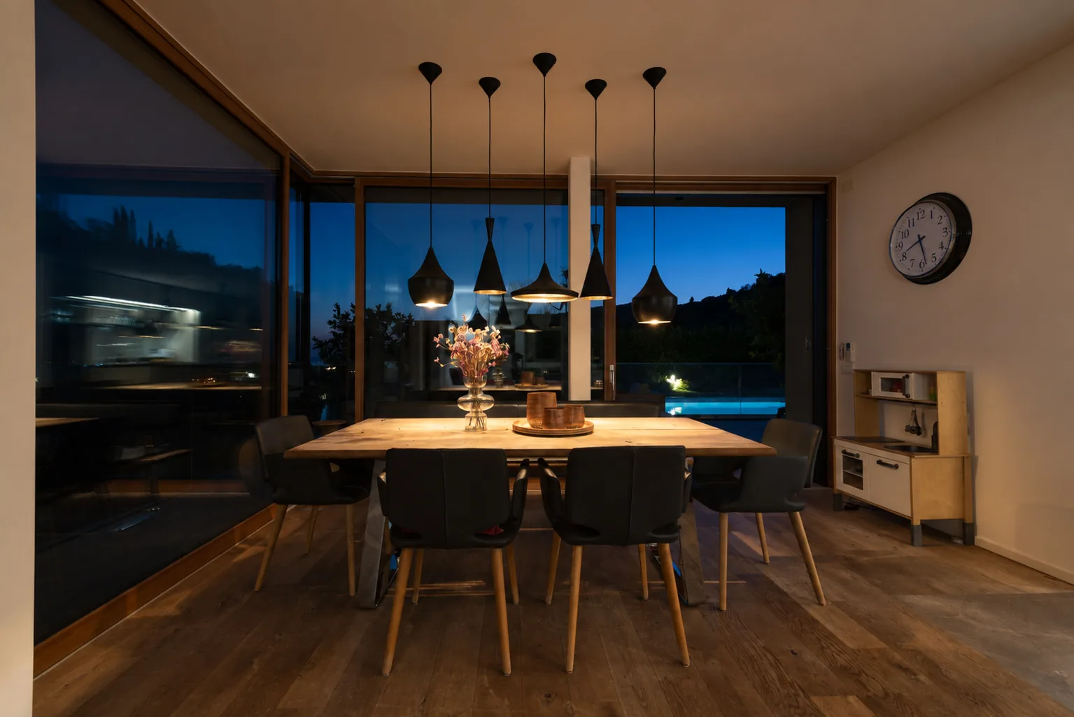 Dining table lit by pendant lights at night, with the blue evening sky visible through large windows. | Only on SopranoVillas